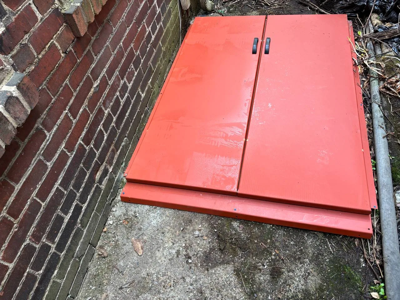 Red metal cellar door beside a brick exterior wall.